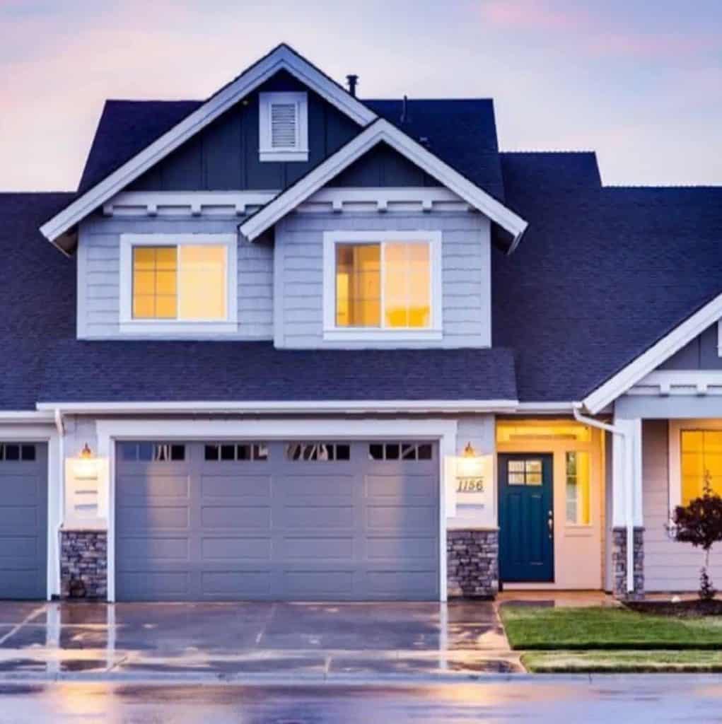 Modern two-story house with gray siding, a double garage, blue front door, and illuminated windows, photographed at dusk with a wet driveway.