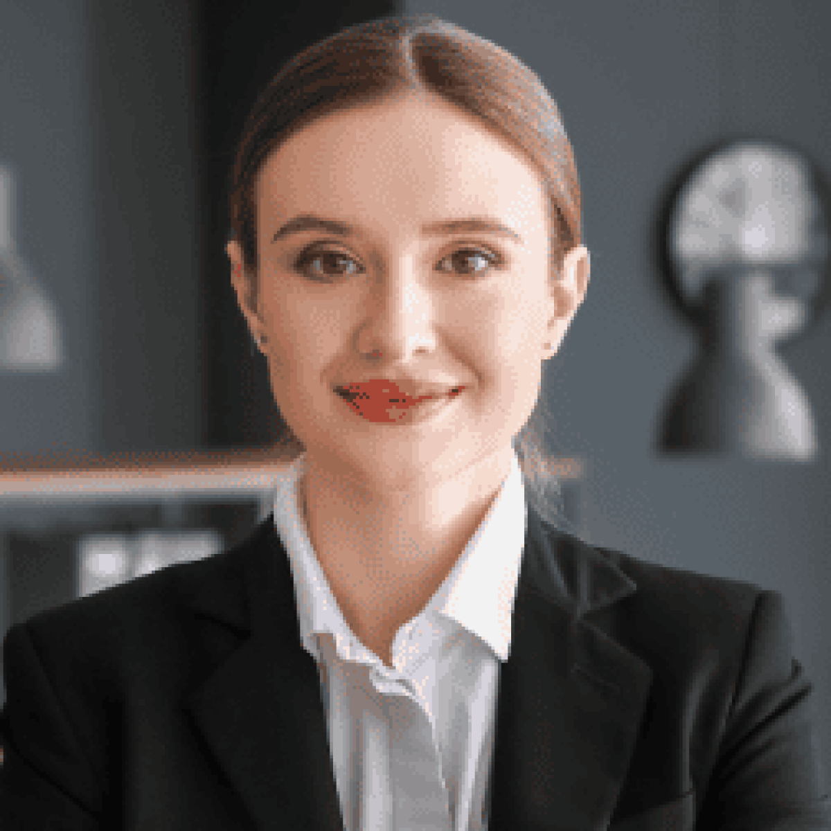 A person in a black blazer and white shirt smiles confidently in an office setting with gray walls and modern lighting.